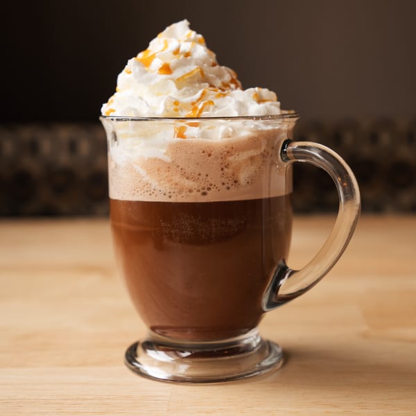 A clear mug of coffee with whipped cream and caramel drizzle sits on a light wooden surface.