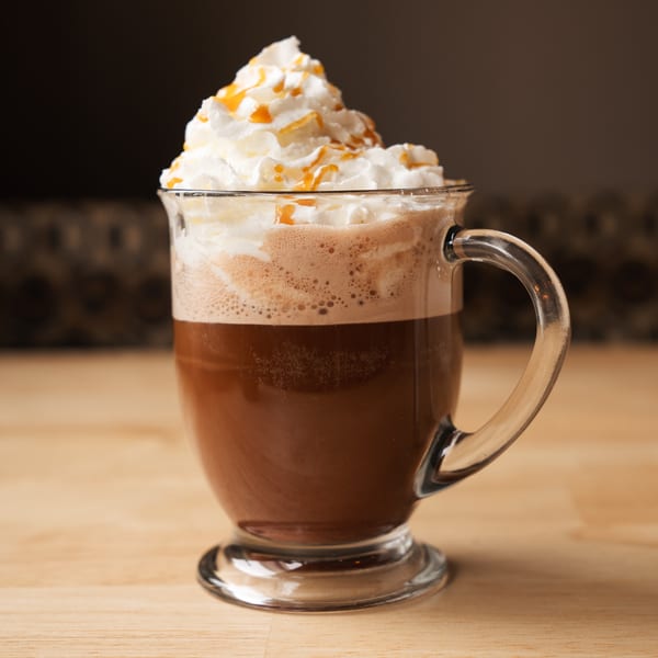 A clear mug of coffee topped with whipped cream and caramel drizzle on a wooden surface.