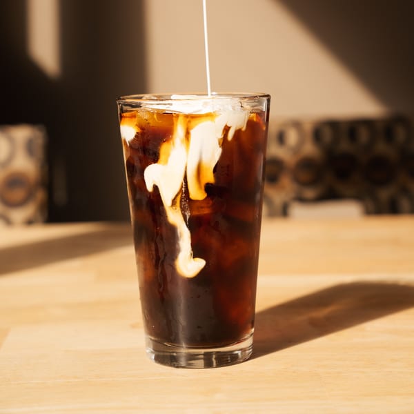 Creamy milk being poured into a tall glass of iced coffee on a wooden table.
