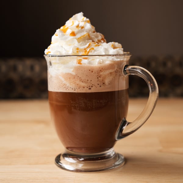 A glass mug of coffee topped with whipped cream and caramel drizzle on a wooden table.