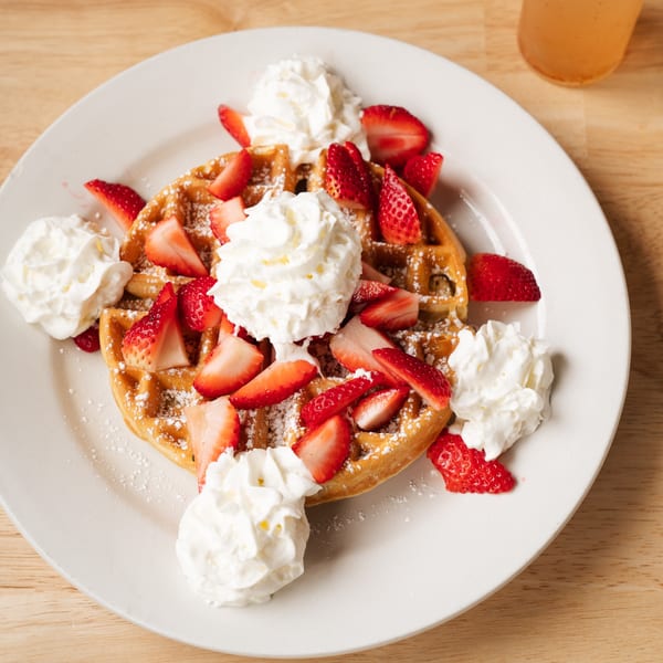 Golden waffle topped with fresh strawberries, whipped cream, and powdered sugar on a white plate.