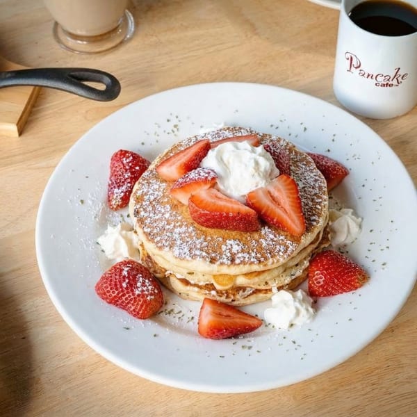 Stack of Pancake Cafe pancakes with strawberries, whipped cream, and powdered sugar, next to coffee.