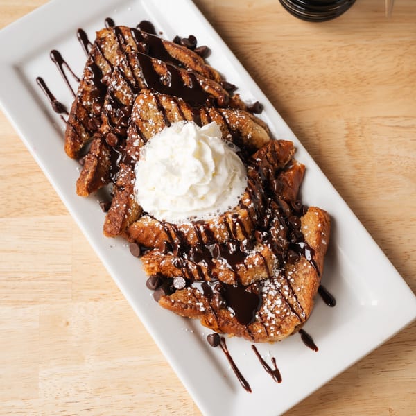 Chocolate chip French toast with whipped cream, chocolate drizzle, and powdered sugar on a white plate.