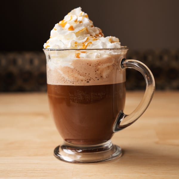 A glass mug of coffee with whipped cream and caramel drizzle on a wooden table.