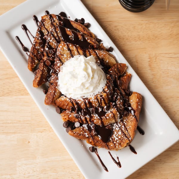 Chocolate chip french toast with whipped cream, chocolate drizzle, and powdered sugar on a white plate.