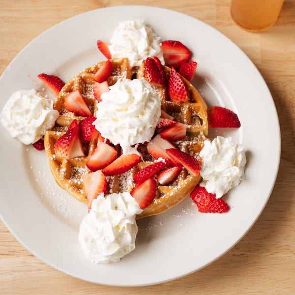 A golden waffle topped with sliced strawberries, whipped cream, and powdered sugar on a white plate.
