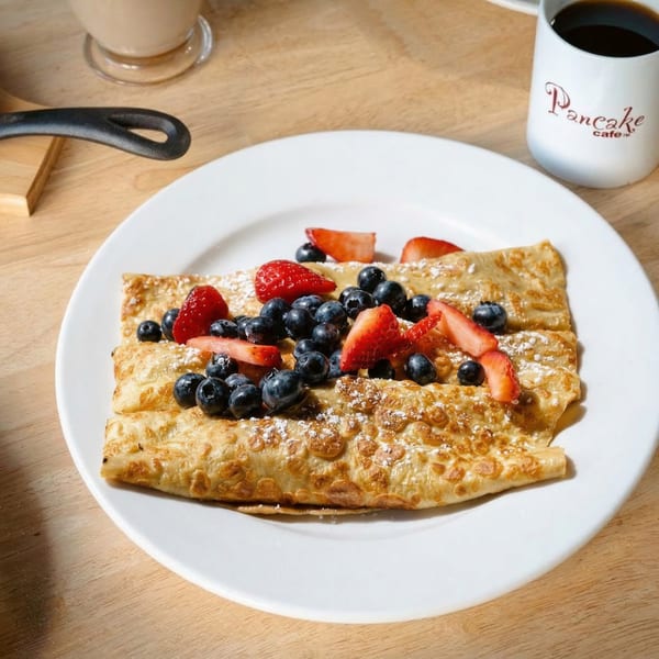 Pancake Cafe crepes with blueberries, strawberries, and powdered sugar, next to a coffee mug.