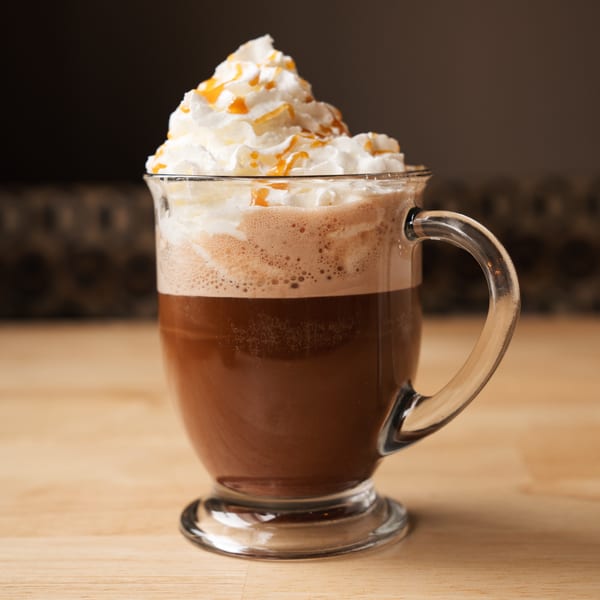 A glass mug of coffee topped with whipped cream and caramel drizzle on a wooden surface.